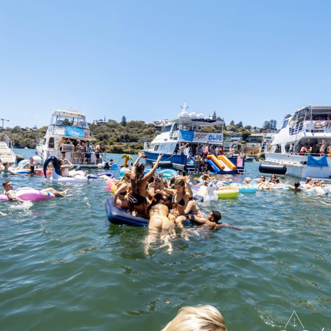 A lively scene of people enjoying a sunny day on the water. Groups of friends are on colorful inflatables and boats, socializing and having fun. The background features several anchored boats and a picturesque shoreline with trees. The atmosphere is vibrant and relaxed.