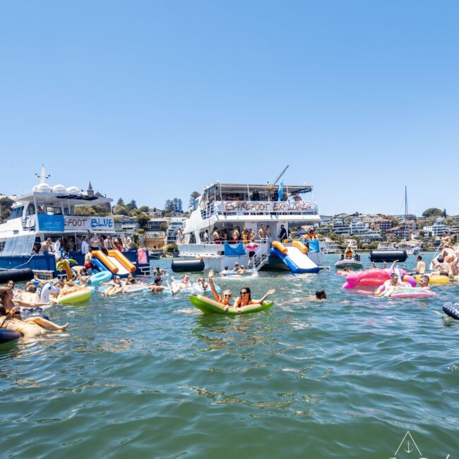 A lively scene of people enjoying a sunny day on the water. Many are floating on various inflatable rafts, socializing and having fun. Several yachts are anchored nearby, with more people on board. In the background are tree-covered hills and houses. "Yacht Social Club" is seen in the bottom right corner.
