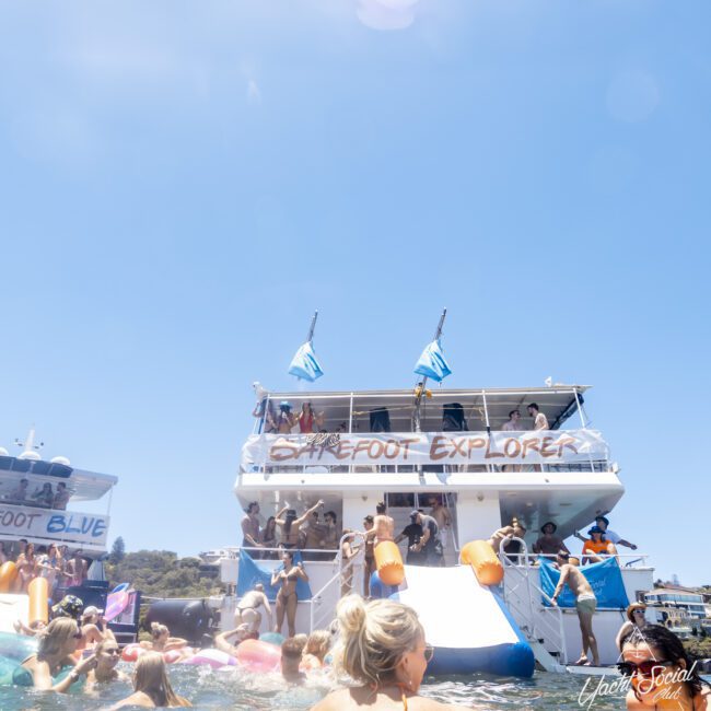 A vibrant scene of people enjoying a sunny day on a large boat named "Shoes Feet Explorer." Decorated with flags, the boat has people swimming and lounging around it. Colorful inflatable floats in the water enhance the festive atmosphere, making it perfect for relaxation and fun.