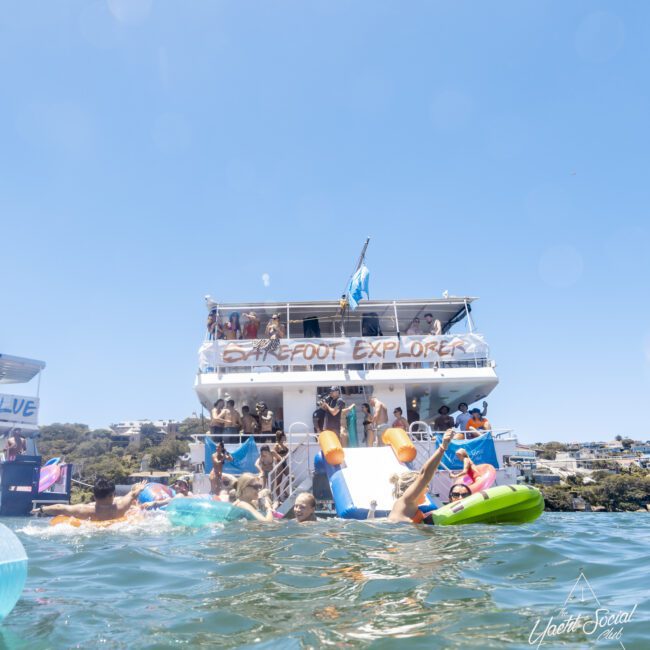 A double-decker boat named "Barefoot Explorer" on the water is crowded with people enjoying a sunny day. Some people are swimming or floating on colorful paddle boards nearby, and the lively scene features a backdrop of trees and buildings. A logo saying "Yacht Social Club" is in the lower right.
