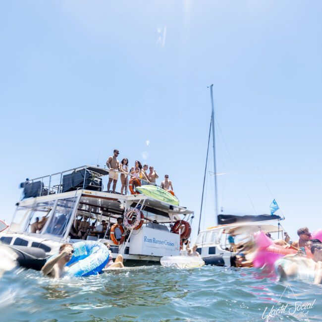 A group of people enjoying a lively party on a large two-level boat named "Rum Runner Cruise." Some are standing on the upper deck under a canopy, while others float in the water on inflatables. Another boat and splashing water are visible in the background. Text reads "Yacht Social.