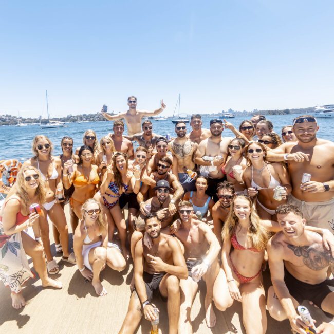 A large group of people in swimwear pose together on a yacht on a sunny day. The background shows calm waters with a few other boats and a distant shoreline. The group appears cheerful and is enjoying drinks, with some people standing and others seated, capturing the essence of summertime fun.