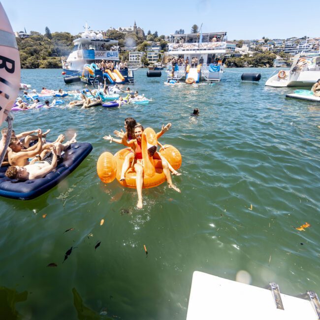 People are enjoying a sunny day on a lake with various inflatables and boats in the background. A group of friends is on an orange inflatable in the foreground while others relax on blue floats. The shore has several charming houses and lush greenery, creating an inviting atmosphere for all.
