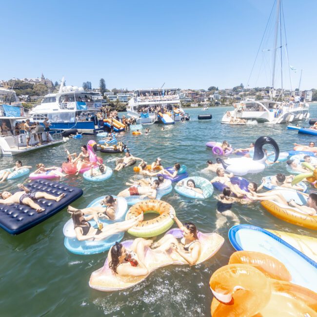 A lively scene with numerous people relaxing on colorful inflatable floaties in a bustling body of water, surrounded by several yachts. The floaties vary in shapes and sizes, and the atmosphere appears festive and leisurely under a bright, clear sky.
