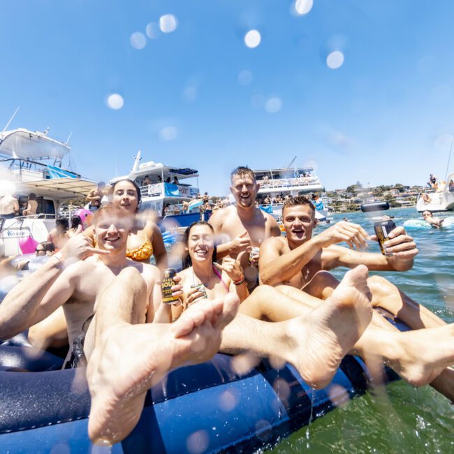 A group of smiling young adults sit on an inflatable raft in the water, holding drinks. They are surrounded by boats under clear blue skies. Water droplets create a lively effect in the foreground. Everyone is in swimsuits, enjoying a sunny and carefree day with music playing nearby.