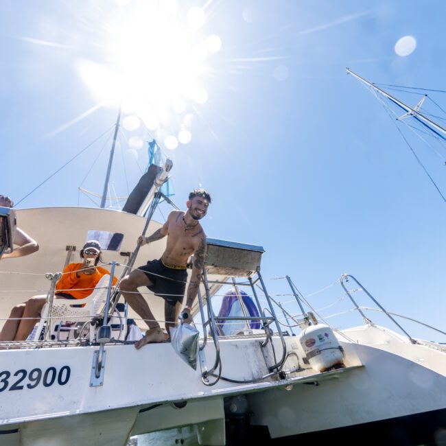 A shirtless man stands on a docked boat's railing, holding onto the ladder under the bright sun. Other people are visible on the boat marked "82900." The calm, green water below contrasts with the clear blue sky above.