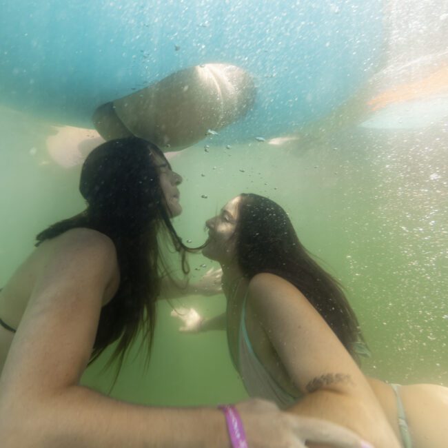 Two women are underwater, holding hands and facing each other with smiles. They appear to be beneath an inflatable raft or boat, which is visible above them. The water is slightly murky, giving a greenish tint to the scene, adding a mystical touch to their joyful encounter.