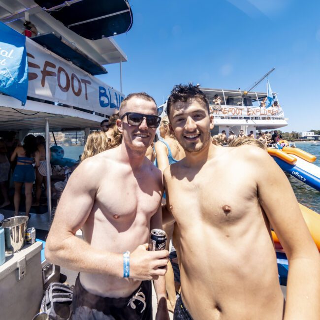 Two shirtless men smile at the camera on a boat crowded with people and inflatable pool toys. They are enjoying a sunny day by the water, with a sign in the background reading "Barefoot Social Club." Other partygoers are visible, soaking up the fun.