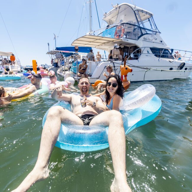 A man and a woman relax on a blue inflatable lounger in the water, both smiling. Behind them, several boats with people are anchored. Other people are also seen floating nearby on inflatables under a clear, zen sky. The scene appears festive and joyful.