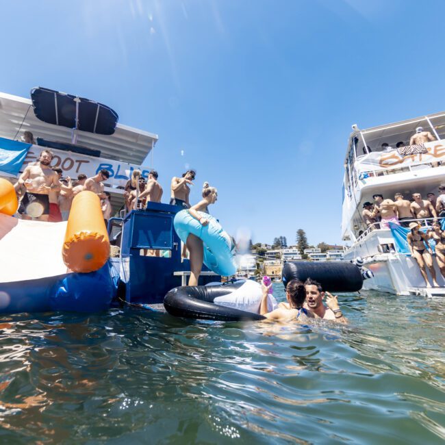 A lively scene on the water with two large boats filled with people partying. Some are dancing, and others are using inflatable slides and tubes to splash into the water. One boat has "Seafoot" written on it, while kayakers paddle nearby. The sky is clear, and the atmosphere is festive.