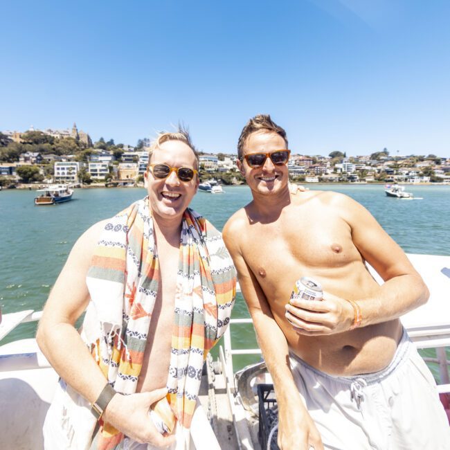 Two men smiling and enjoying a sunny day on a boat with a scenic coastal backdrop. One is holding a drink, wearing sunglasses and has a colorful towel draped over his shoulder, while the other is shirtless, wearing sunglasses and holding a drink. Other joyful people are on the boat.