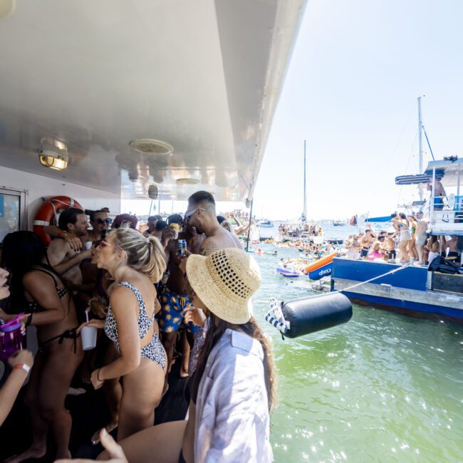 A group of people in swimsuits gathers on the deck of a boat, enjoying a sunny day on the water. Another boat with more people is docked nearby. Some individuals are in the water, and the atmosphere appears festive and social, perfect for celebrating with friends.