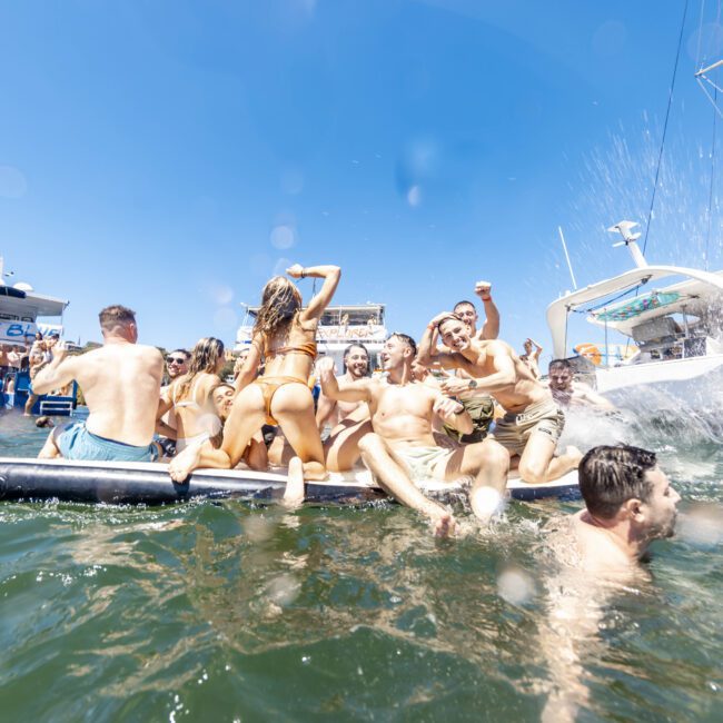 A lively group of people in swimsuits enjoy a sunny day on an inflatable platform in the water, basking under the clear sky. More people are swimming nearby, with several yachts anchored in the background. The atmosphere is festive, with one person raising their arm in celebration.