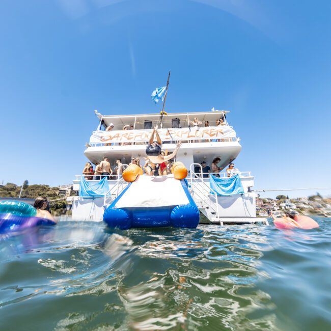 A large yacht named "Caribbean Explorer" is anchored as people swim around it in clear blue water. There is a slide from the yacht's upper deck into the water. The beautifully adorned vessel sets the scene for several people enjoying exhilarating water-based activities.