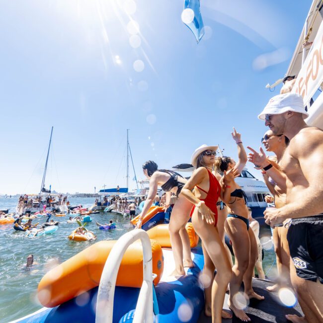 A lively group of people in swimsuits and summer hats is gathered on a boat deck, ready to jump into the water. Numerous others are swimming, floating on colorful inflatable tubes, and soaking up the sun. Multiple boats are anchored in the background under a clear blue sky, creating a perfect summer scene.