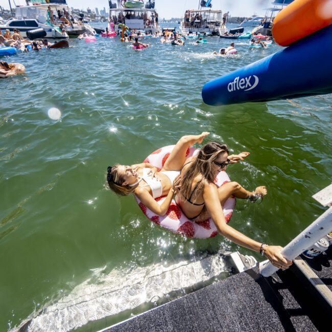 A lively scene of people enjoying the water near several anchored boats. Two women float on an inflatable donut ring by a slide extending from a dock. Many others are swimming, playing, or lounging around, creating a festive and relaxed atmosphere under the sunshine.