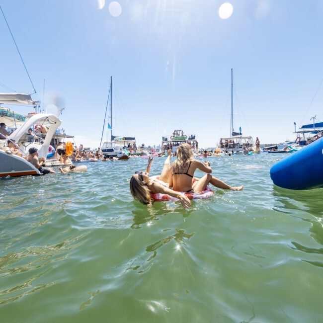 A vibrant scene of a yacht social event on a sunny day. Boats are gathered in a calm, green-blue ocean, with people socializing on board and enjoying the scenic coastline. In the foreground, two women are relaxing on a pink inflatable float in the water.
