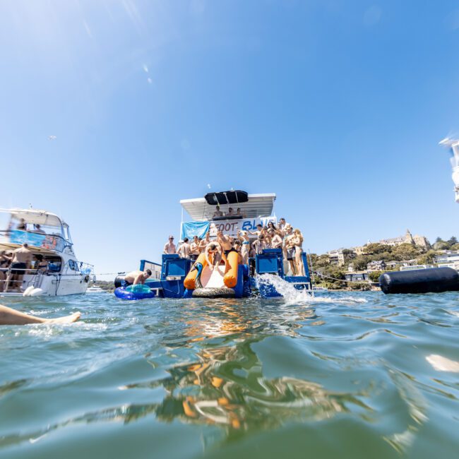 A group of people are gathered on the deck of a boat, having a lively party. Some are swimming in the water nearby, and other boats with people are visible in the background. The scene is bright and sunny, showcasing summer fun. The boat is anchored in a scenic spot with a clear, blue sky.