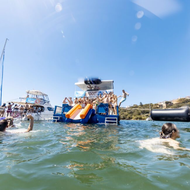 People are on boats enjoying the water on a sunny day. Some are using a waterslide from one of the boats, and others are swimming nearby. Several boats are anchored together, creating a lively and festive summer atmosphere perfect for water sports enthusiasts.