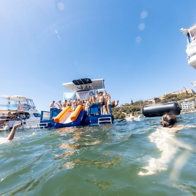 A group of people on boats and floating in the water, some enjoying orange water slides from the boats. The sky is clear and blue, with people both in the water and on the decks of the boats having fun in the sun. A logo is visible in the bottom right corner.