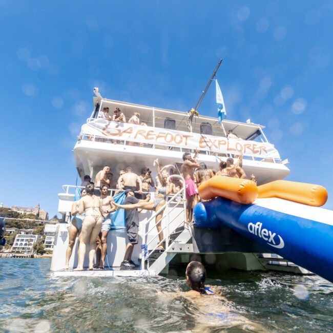 A lively scene on the deck of a boat named "Barefoot Explorer." People are enjoying themselves, with some eager to slide down the bright blue and orange waterslide into the shimmering water. The sun is shining beautifully against a clear blue sky.