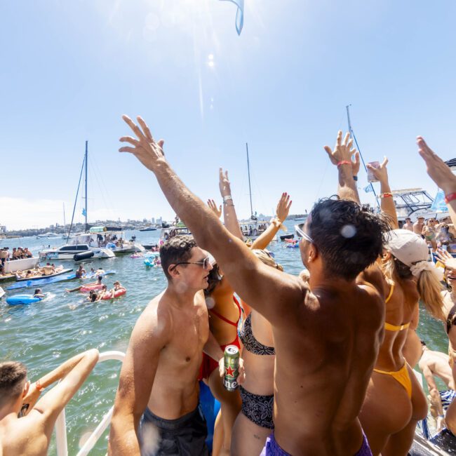 A lively crowd in swimwear celebrates on a boat under clear blue skies. Arms are raised, drinks in hand, as others swim and float on the water nearby. Boats are in the background, contributing to the vibrant, summer atmosphere by the beach.