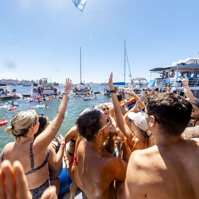 A lively group of people enjoying a sunny day on a boat, some raising their hands and smiling. The background shows several more boats and people swimming in the water. The festive atmosphere is full of vibrant energy. The logo "Yacht Social Club" is visible in the bottom right corner, adding to the scenic vibe.