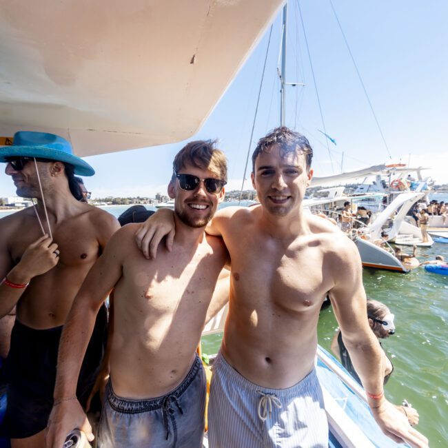 Four shirtless men enjoy a lively social event on a boat. One, wearing sunglasses, embraces another in the foreground. The third, sporting a blue hat, stands to the left while the fourth is blurred in the background. The water teems with boats and people, adding to the festive atmosphere.