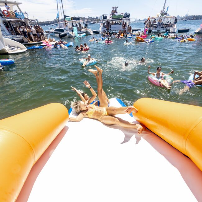 People are enjoying a lively boat party on the water under a sunny sky. Several individuals are swimming and lounging on inflatable floaties near numerous boats. In the foreground, a person is sliding down an enormous inflatable slide into the refreshing water.