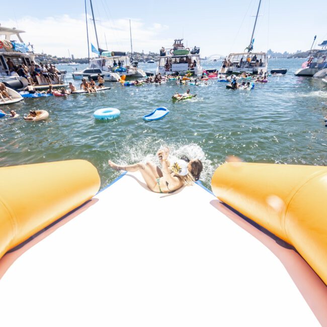 A crowded, sunny beach scene with numerous anchored boats and people swimming or floating on inflatables in the water. In the foreground, a person splashes into the water from an inflatable slide attached to a boat. The relaxing vibe is enhanced by the "Check Social Club" logo in the bottom right corner.