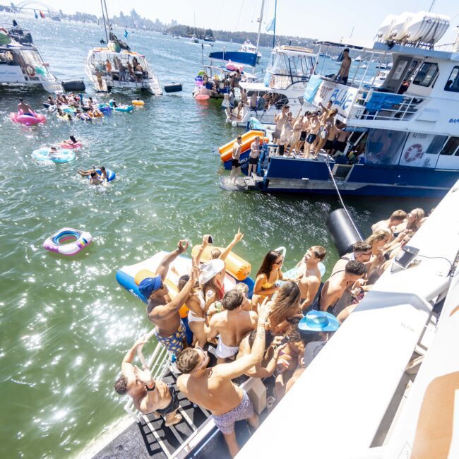 A lively boat party is in progress on a sunny day. Many people in swimsuits are gathered on the deck of a boat, socializing, dancing, and drinking. Several other boats surround the area, and some partygoers are paddleboarding. The scene is joyful and energetic.