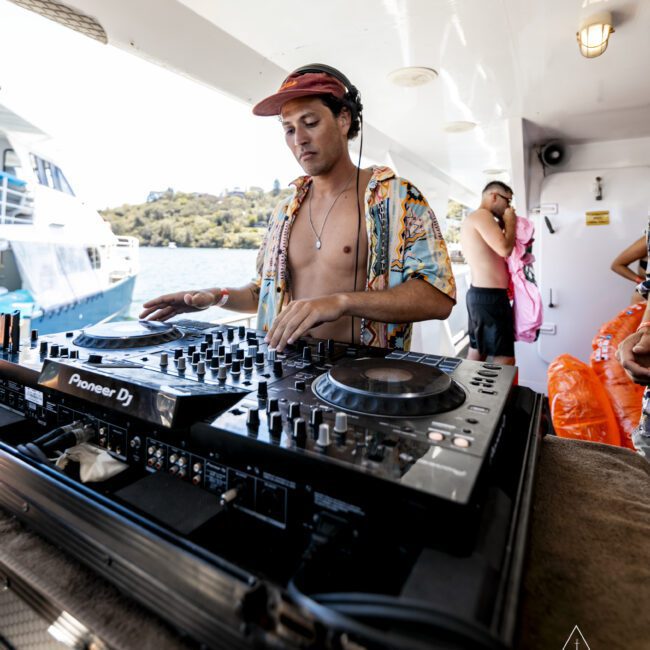 A DJ stands behind a mixing console on a yacht, wearing a floral patterned shirt that is unbuttoned and a red cap. Another person in the background, shirtless and wearing sunglasses, stands near the railing. A jet ski zooms by in the sunny scene with water and another boat visible.