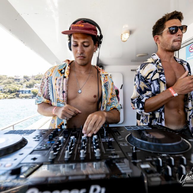 Two men in vibrant shirts operate DJ equipment on a boat under bright sunlight. The man on the left wears a hat and headphones, focused on the mixer, while the other stands with sunglasses and a drink, enjoying the energetic atmosphere. Water and distant land are visible in the background.