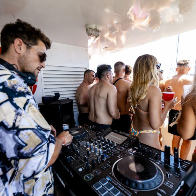 A DJ stands behind a mixer and turntables, spinning music at a party. The scene is crowded with people in swimwear, enjoying drinks and dancing. A vibrant, sunny, beach-like atmosphere is visible with a lifebuoy attached to the wall in the background, enhancing the festive vibe.