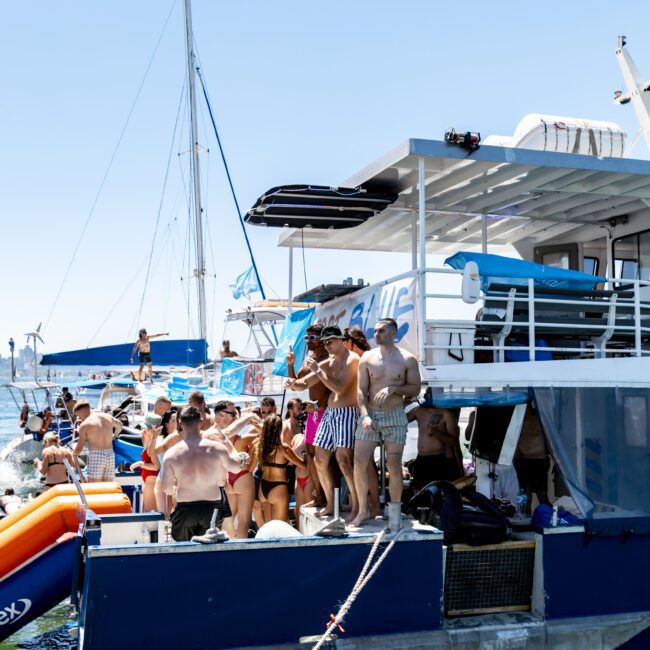 A large group of people, many in swimsuits, gather on a docked yacht. Several inflatable pool floats are nearby, and other boats are visible in the background on the water. The scene is sunny and festive, suggesting a lively social event on the yacht.
