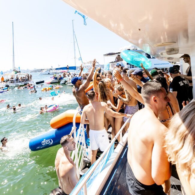 A lively boat party is underway with people dancing and having fun on a docked boat, while others enjoy the water using inflatables and swimming. The bright sunny day and clear blue sky add to the festive atmosphere, with various colorful floatation devices adding to the scene.