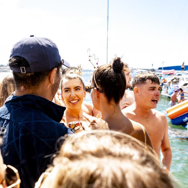 A group of people in swimwear are gathered and chatting at a lively boat party on a sunny day. One woman with a bright smile and decorative headband stands out among the crowd. In the background, other partygoers enjoy themselves aboard nearby yachts. The bottom right corner has a logo that reads "Yacht Social Club.