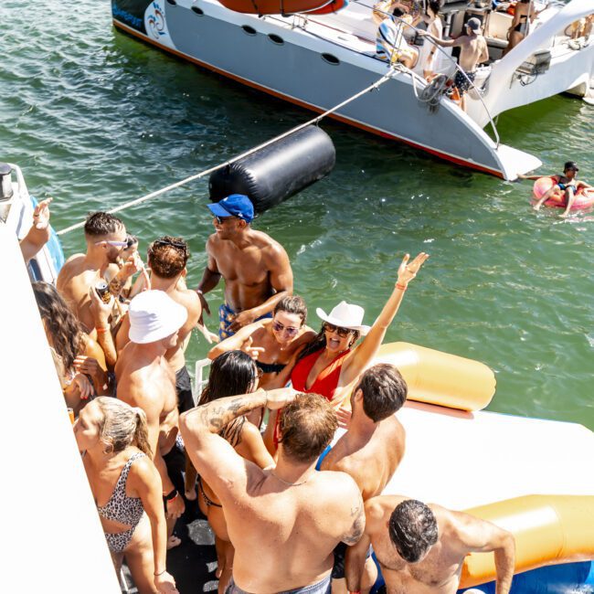 A group of people in swimwear are gathered on the deck of a boat, dancing and cheering under sunny skies. Other boats with more people can be seen in the background on the water. Some attendees are wearing hats and shades, and the atmosphere is lively and festive.