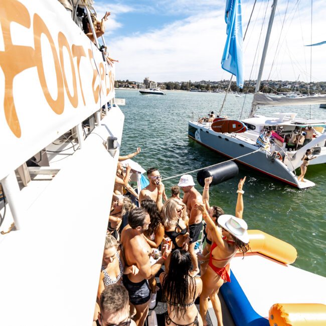 A lively group of people in swimwear are partying on a yacht. Some are dancing and raising their hands, while others are gathered near the edge of the boat. The background shows other boats anchored in the water under a sunny blue sky, enhancing the festive atmosphere.