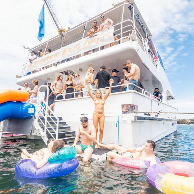 A group of people enjoys a sunny day on a two-story boat named "Barefoot Explorer." Some are on the deck and upper level, while others relax in the water with colorful floaties. A woman stands on the boat's stairs, extending her arms joyfully, as others lounge or swim nearby.