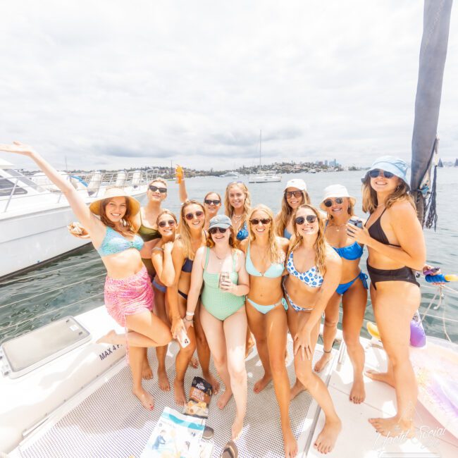 A group of 11 women in stylish swimsuits are gathered on a boat, smiling and posing for a photo. They are enjoying a sunny day on the water, with a city skyline and other boats visible in the background. The mood is cheerful and celebratory, capturing the essence of summer fun.