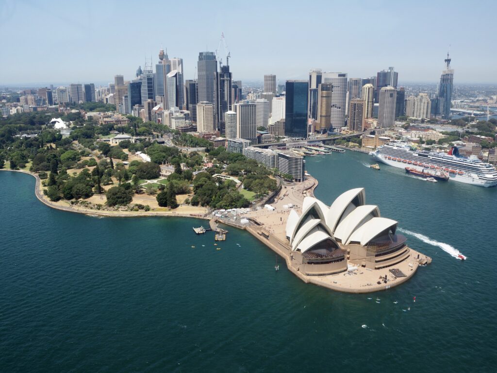Aerial view of Sydney, Australia, featuring the iconic Sydney Opera House surrounded by water. The city skyline with skyscrapers is visible in the background, alongside a large cruise ship docked near the central business district and Bondi Beach in the distance.