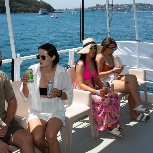 A group of people sit and chat on the deck of a boat under sunny skies. They are drinking canned beverages and wearing casual summer clothing, including swimwear. The background features a calm body of water with several boats and a distant cityscape shimmering against the horizon.