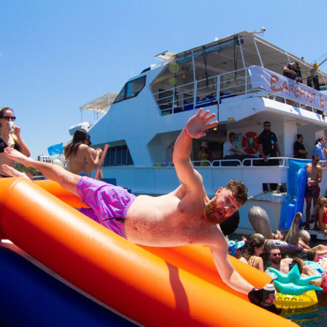 A group of people enjoys a festive boat party. Some are on a large boat, while others are on floats in the water. A man in purple shorts is caught mid-air sliding off an inflated slide into the sparkling blue sea. The atmosphere is lively and celebratory under a clear, sunny sky.