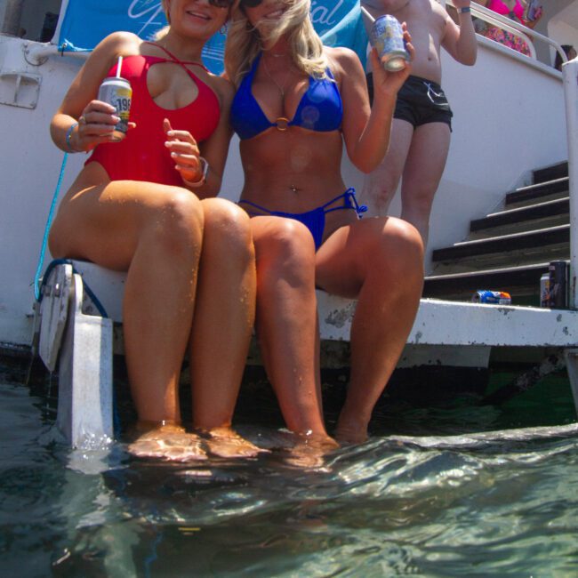 Two women in red and blue swimsuits sit on the edge of a boat with their feet in the water, holding canned drinks. A man in the background also holds a drink, adding to the festive atmosphere under bright sunlight. Other people are visible, enjoying the lively scene.