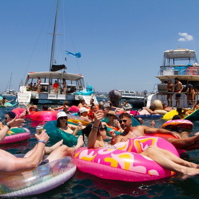 A lively scene of people floating on colorful inflatable pool toys in the water, socializing and enjoying drinks. Boats and yachts are anchored nearby under a clear blue sky, with more people lounging on the decks, creating a festive, relaxed atmosphere reminiscent of a vibrant summer beach party.