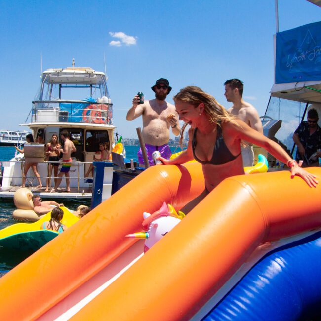 A woman slides down an inflatable slide into the water at a lively boat party. Other people are seen enjoying themselves on and around adjacent boats, some lounging on large floats. The sky is clear, and the atmosphere is festive with laughter and joy.