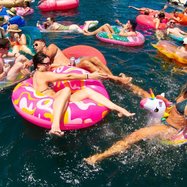 Oriana, Leo, and Angelica are enjoying a sunny day at a pool party, floating in colorful inflatable tubes and a unicorn float. The sparkling blue water is filled with others also relaxing and having fun.