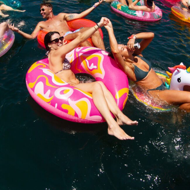 People are floating in a body of water on colorful inflatables, including a pink donut and a unicorn. They are laughing and enjoying the vibes of a sunny day, with the serene landscape adding to their delight.