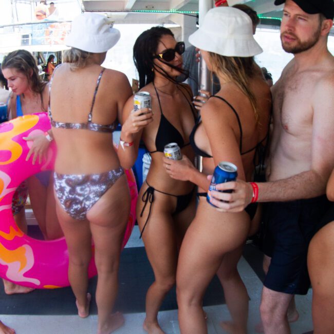 A group of people in swimwear are gathered on a boat. Some hold drinks, and one person has a pink inflatable pool float. They seem to be socializing and enjoying a sunny day with lively music playing in the background. The atmosphere appears lively and relaxed.